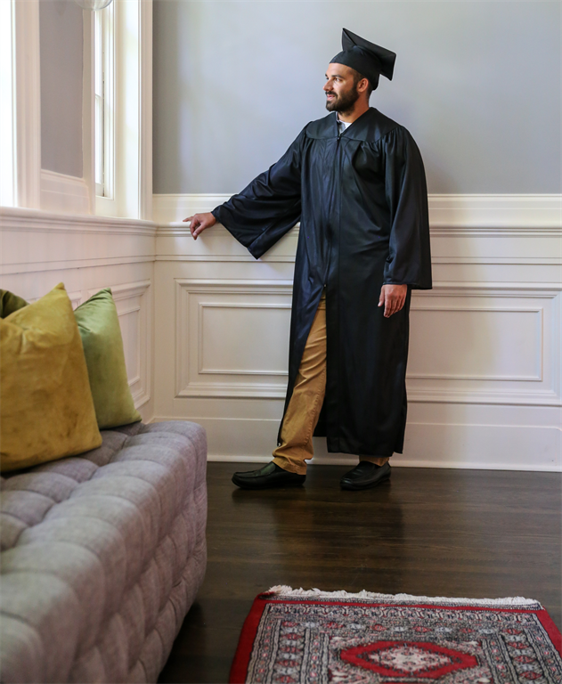man in cap and gown standing on wood floor looking out a window